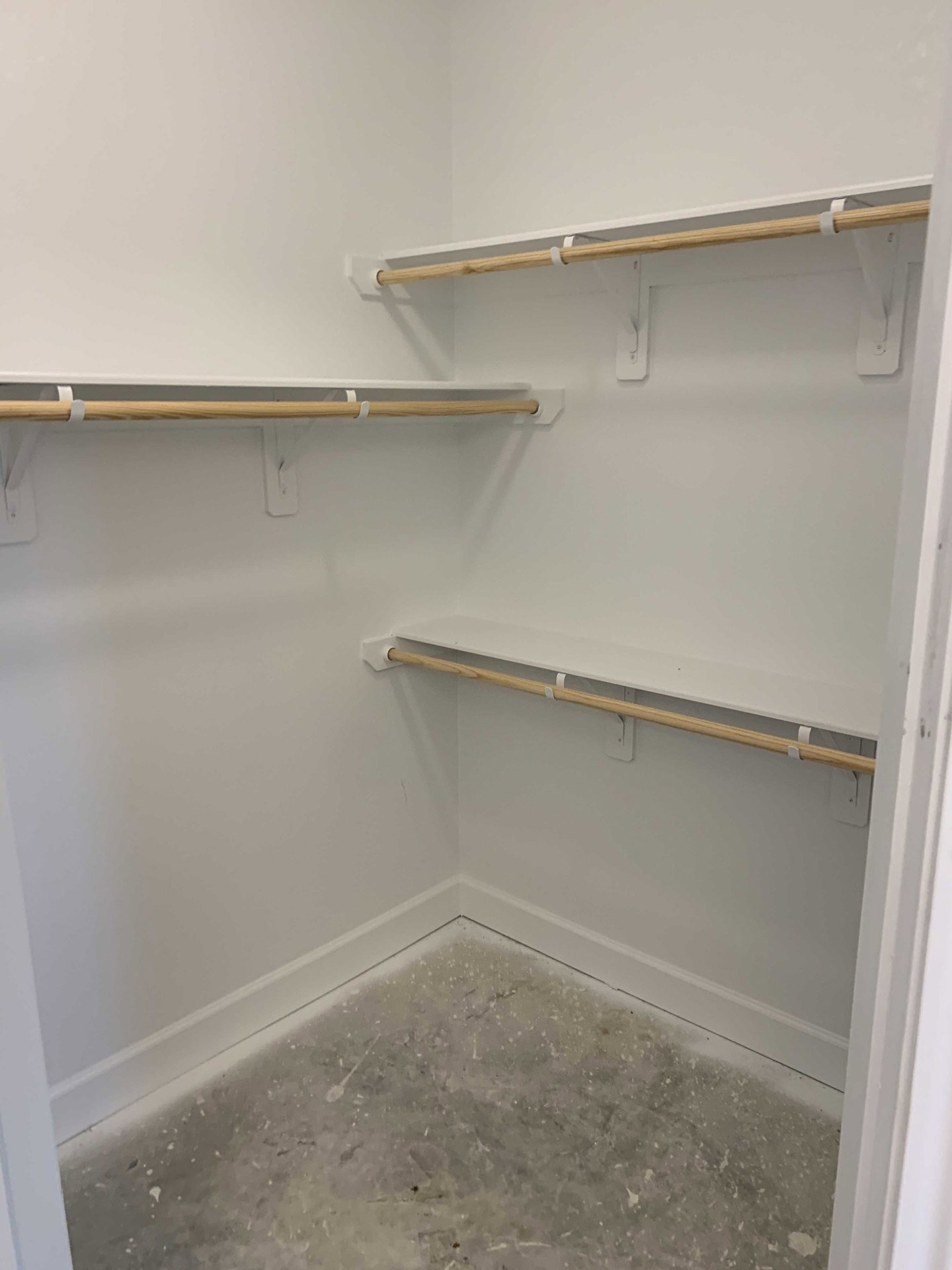 Closet with white shelves, wooden hanging bar, and smooth concrete floor, adjacent to a white plaster wall.