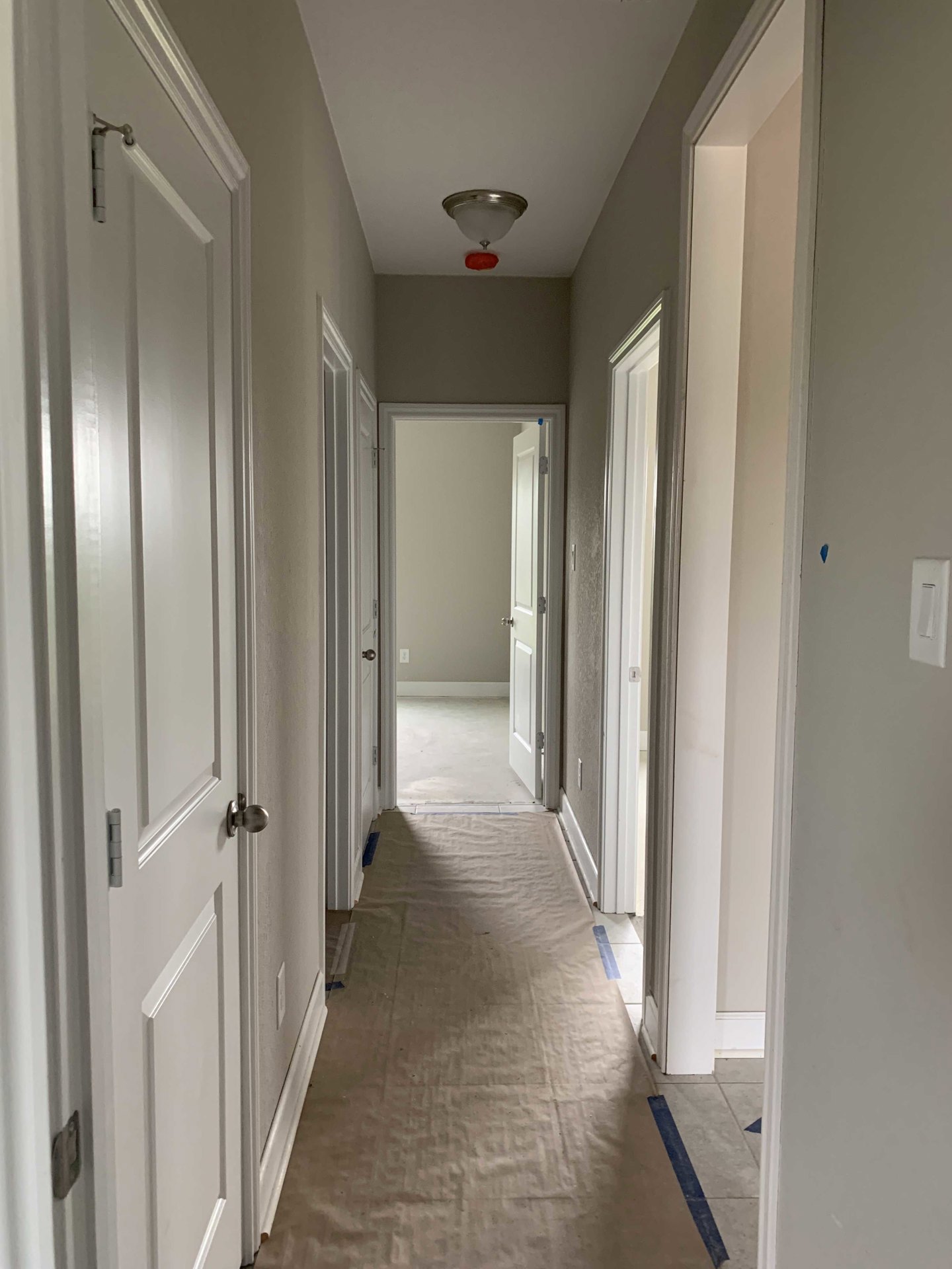 Hallway with smooth white walls, white doors, tile flooring, ceiling light fixture, white curtain with black metal bars, blue painter’s tape on wall, and close-up details of door