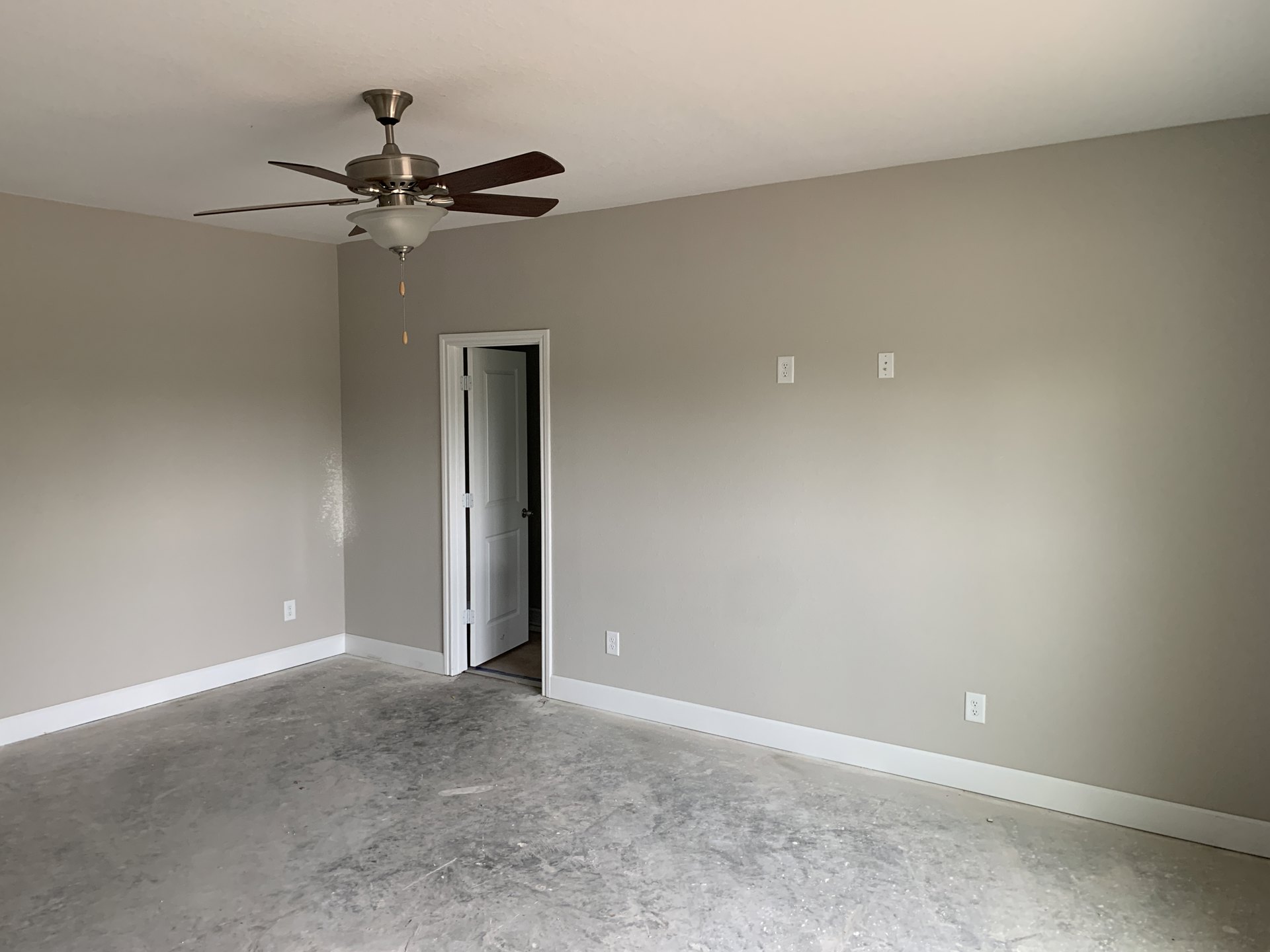 Ceiling fan with integrated light fixture mounted on white plaster ceiling above concrete floor, adjacent to open door with visible hinge.