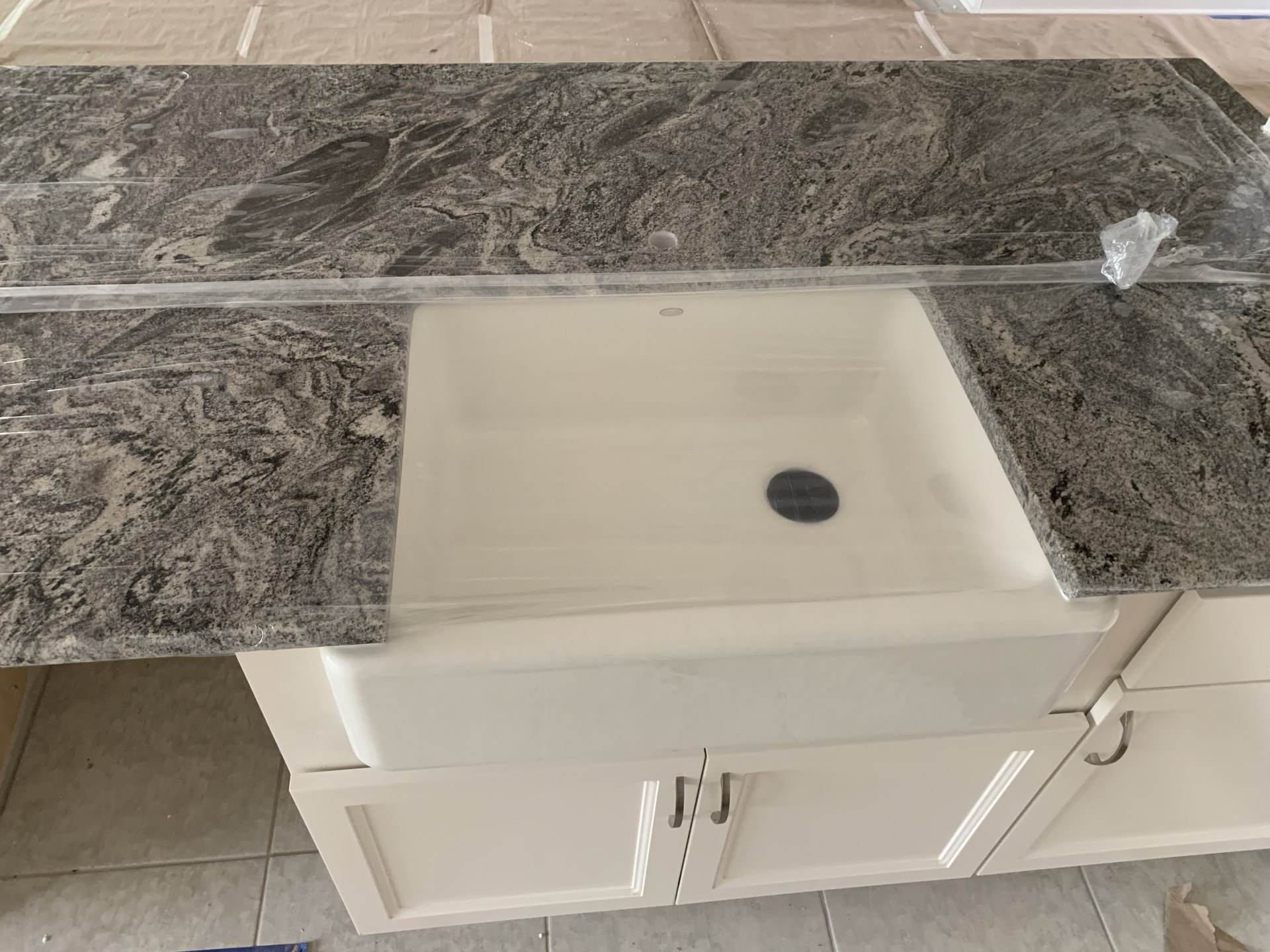 Bathroom with tiled walls and floor, built-in sink on composite countertop, black oval object with white lines, plastic cover over sink, plastic bag on ground, visible plumbing