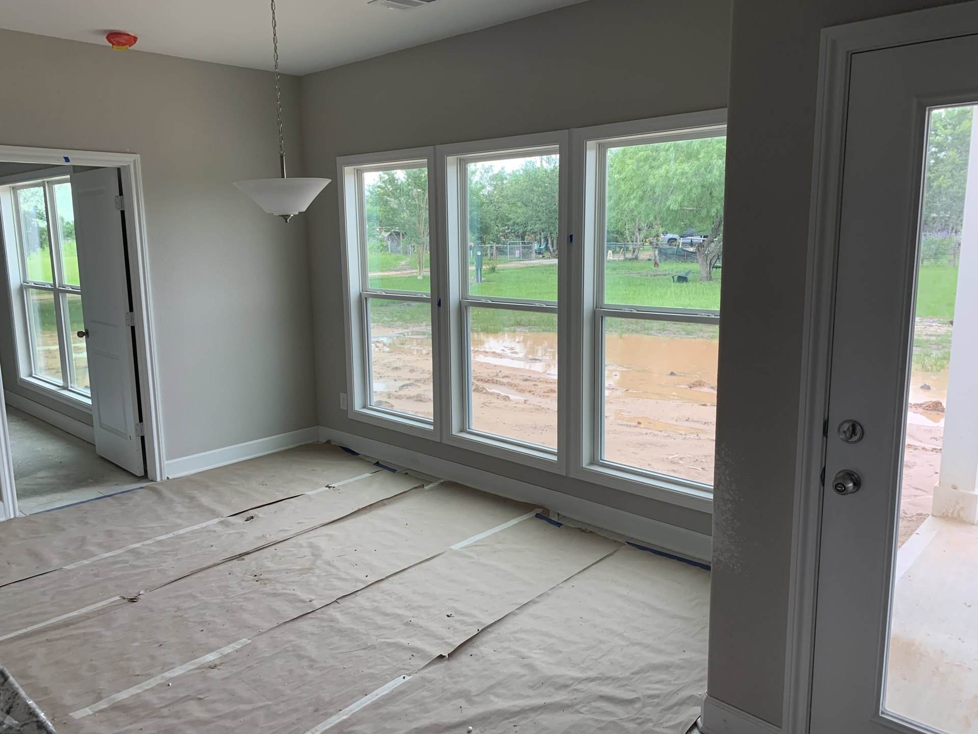 Living room with wide window overlooking flooded yard, open door to outside, paper covering hardwood floor, modern lamp fixture.