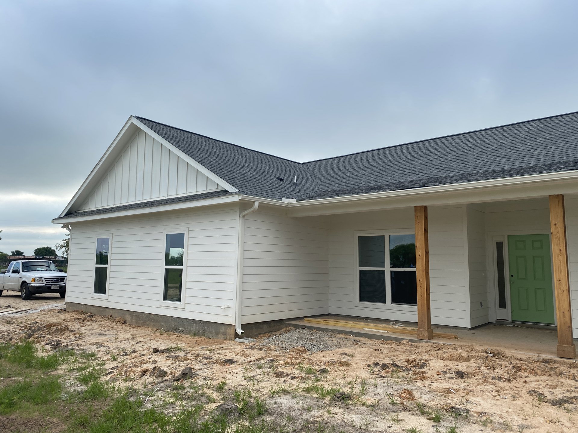 White house with black roof under construction, green front door with black handle, white truck parked on dirt driveway, large windows reflecting trees, cloudy sky overhead
