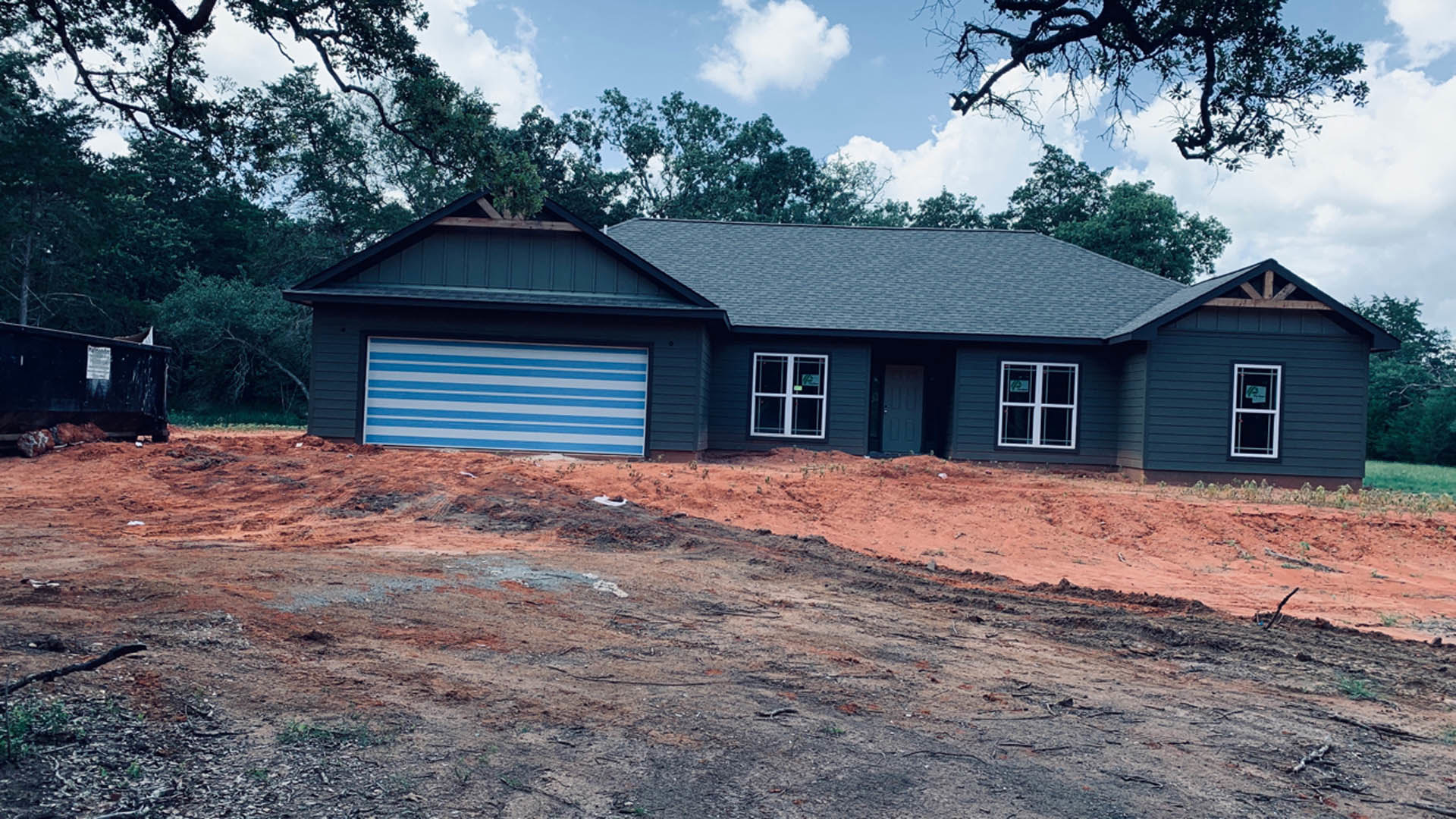Partially built house with blue roof, exposed framing, blue and white striped wall, dirt construction site, dumpster with signage, surrounding trees, and cloudy sky.
