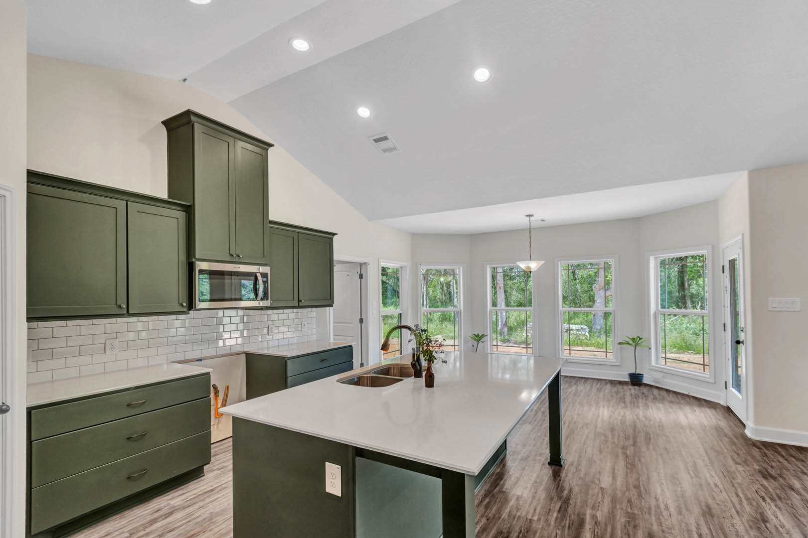 Spacious kitchen featuring a large island with built-in sink and potted plant, green cabinetry with white countertops, wide windows, and stainless steel appliances