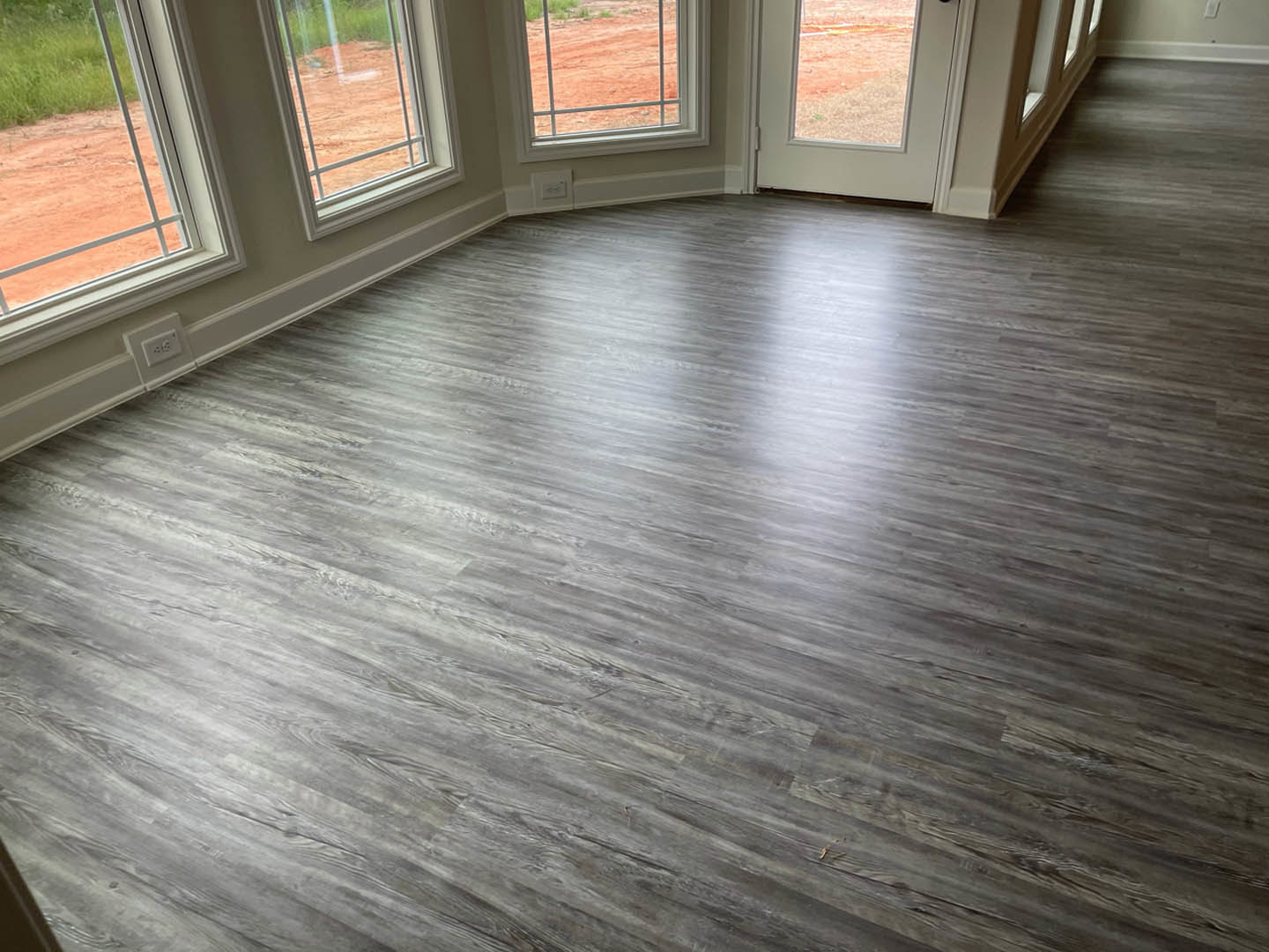 Sunlit room with hardwood plank flooring, white-framed windows overlooking a red dirt field, and a white electrical outlet with black plugs on the wall