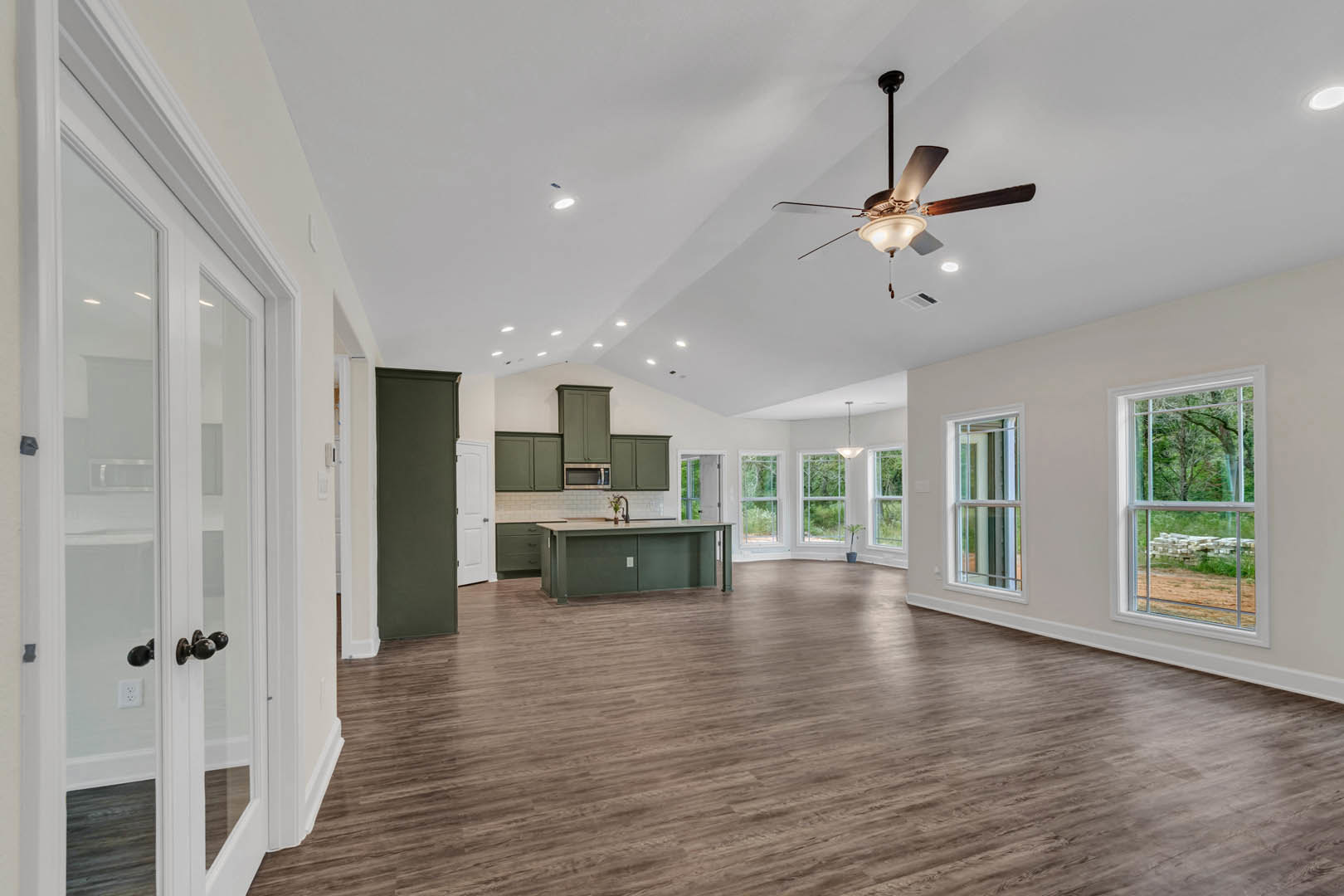 Open-concept living area with wood flooring, ceiling fan with light fixture, green kitchen counter featuring white outlet, large windows overlooking tree and dirt path
