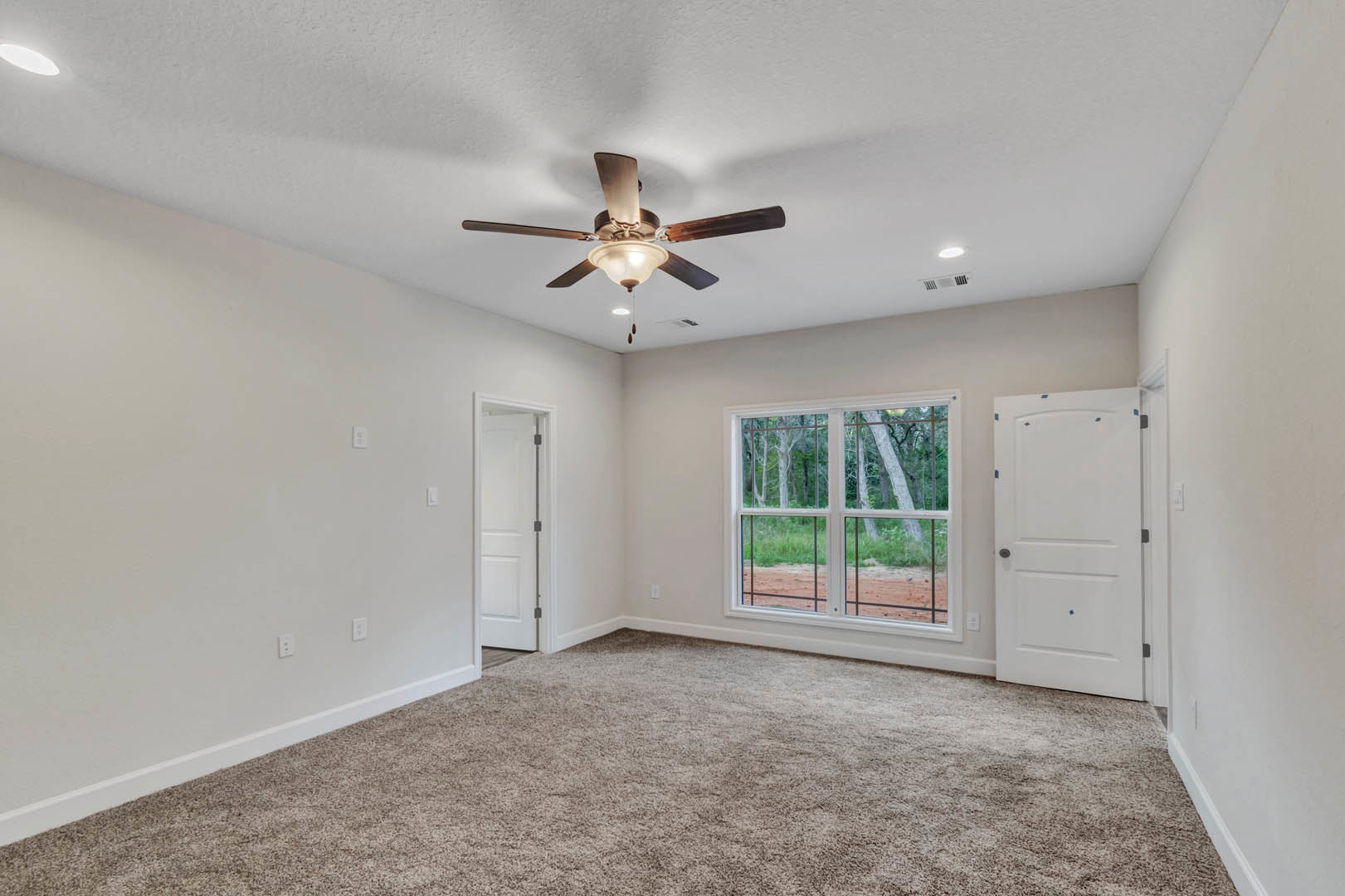 Bedroom with beige carpet, white walls, ceiling fan with light fixture, two large windows overlooking forest, white door with black hardware, crown molding along ceiling.