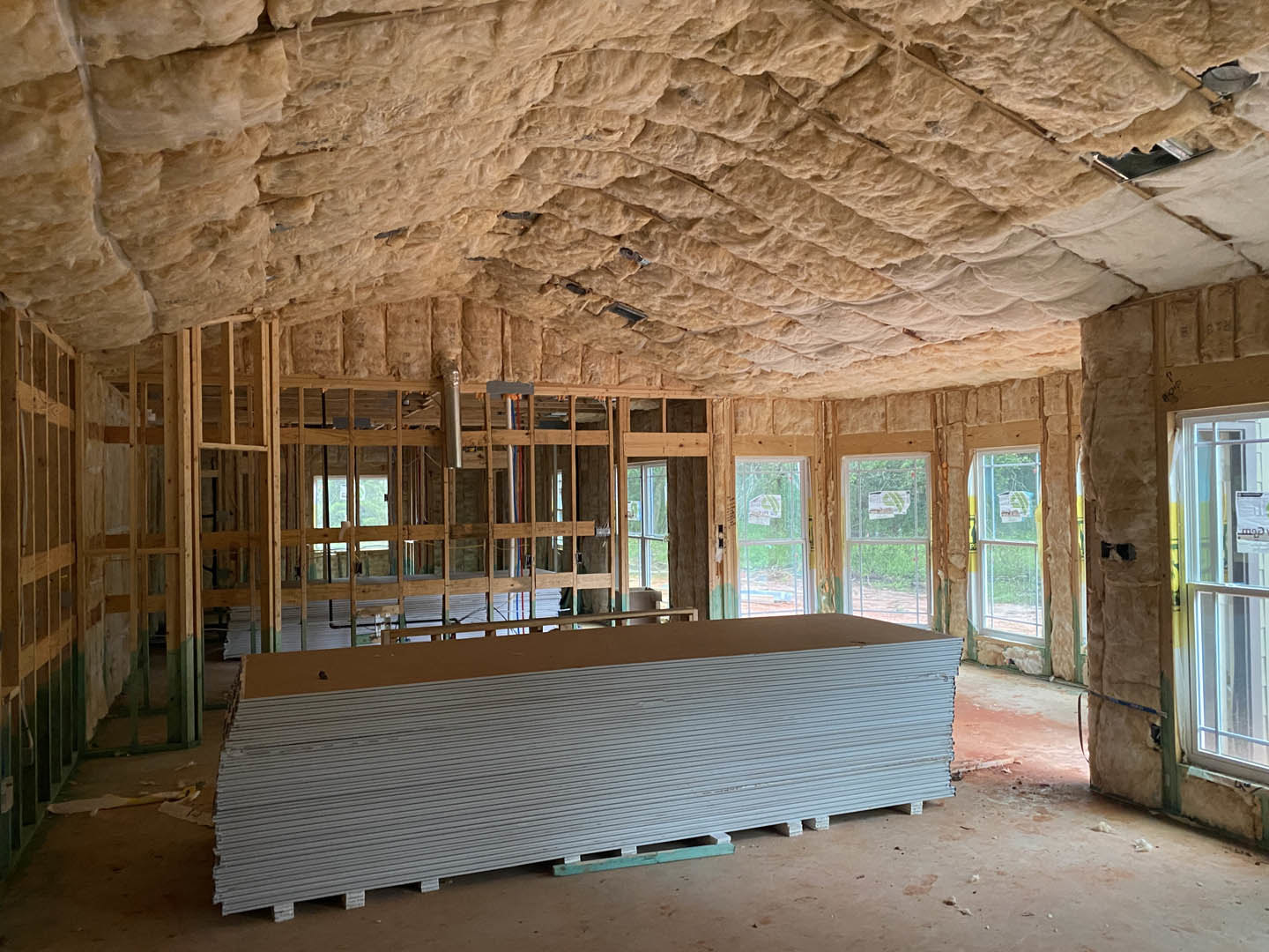 Construction room with stacked plywood sheets, pallet of white boards, insulated ceiling, exposed beams, window displaying a green sign