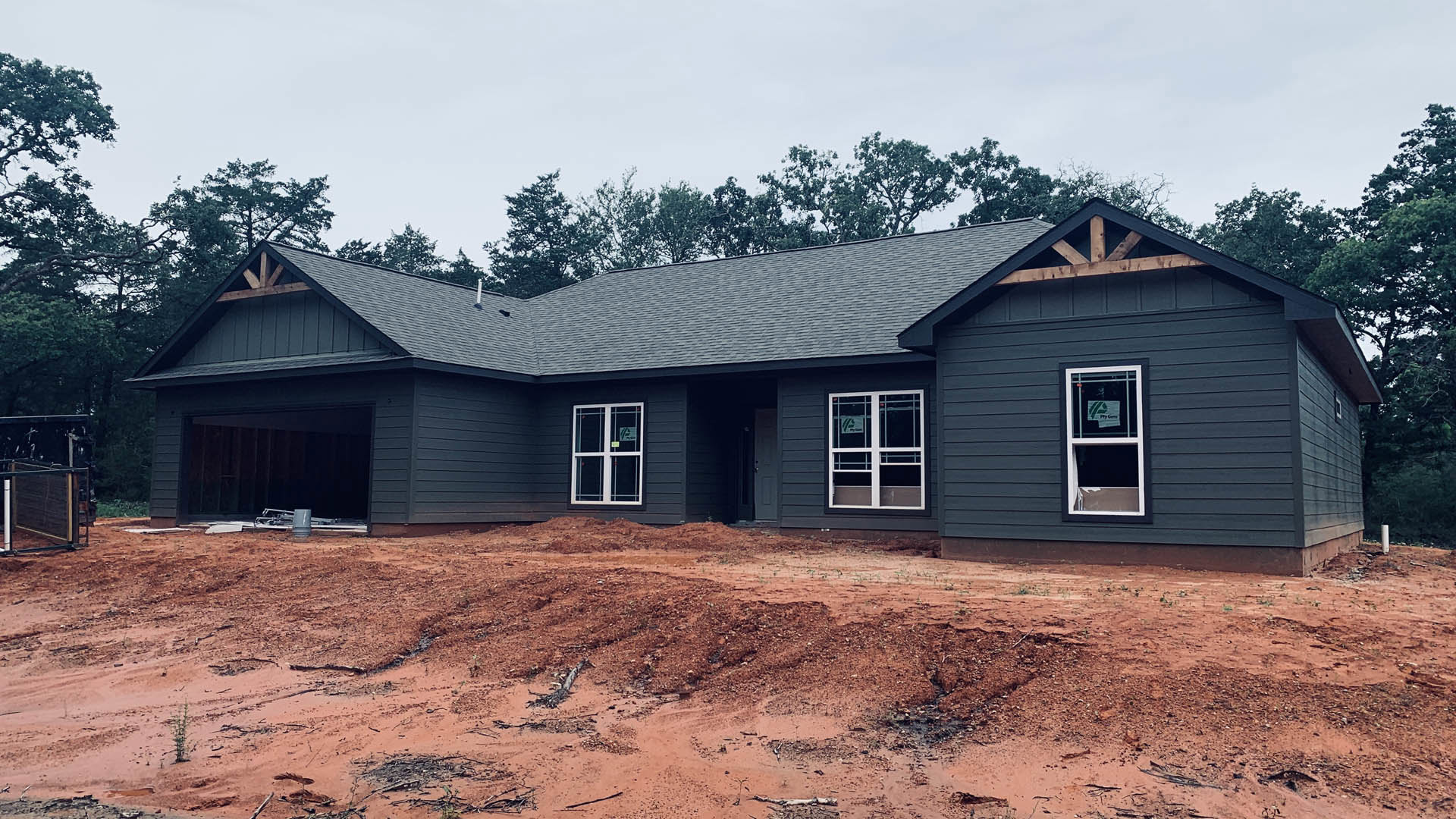 Partially built home with exposed framing, dirt yard, surrounding trees, white door with glass panels, construction sign on window