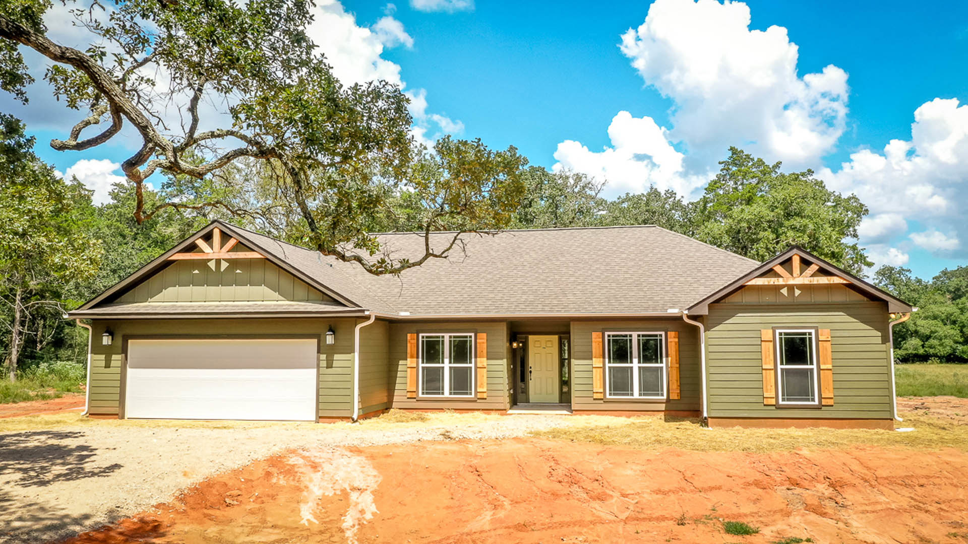 Two-story house with attached garage, white siding, glass-paneled front door, large windows, and dirt driveway bordered by trees under a partly cloudy sky