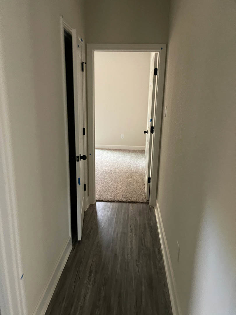 Hallway with dark wood flooring and patterned rug, white door with blue painter’s tape open to carpeted room, plaster walls and ceiling