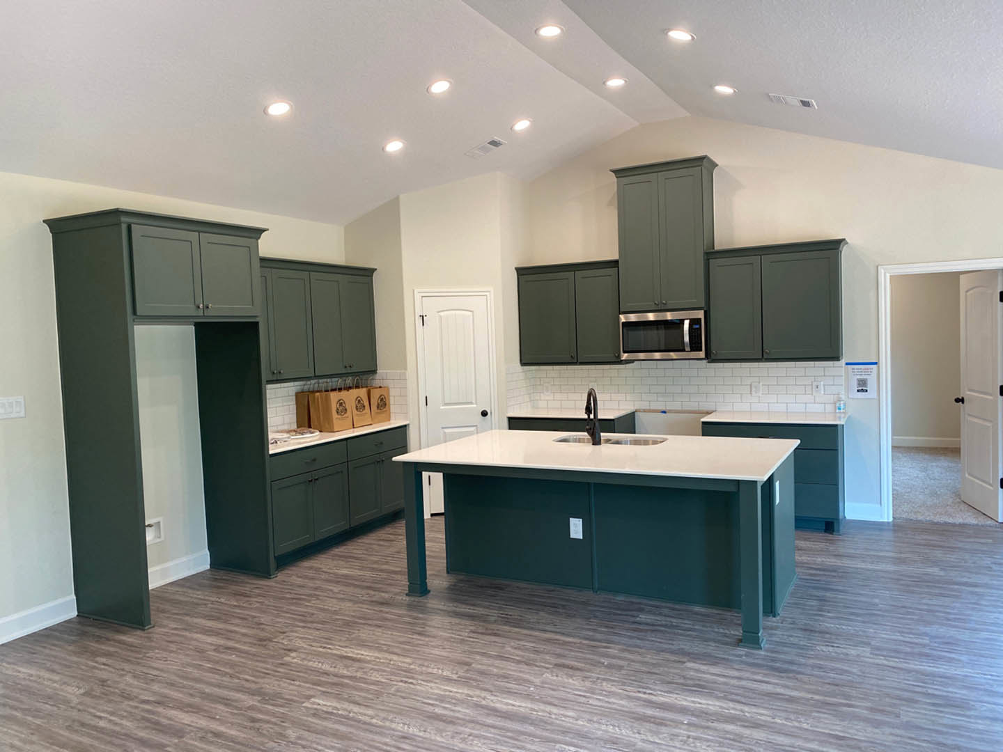 Spacious kitchen featuring a large central island with stone countertop, built-in sink, white cabinetry with black hardware, wood flooring, and green baseboards.