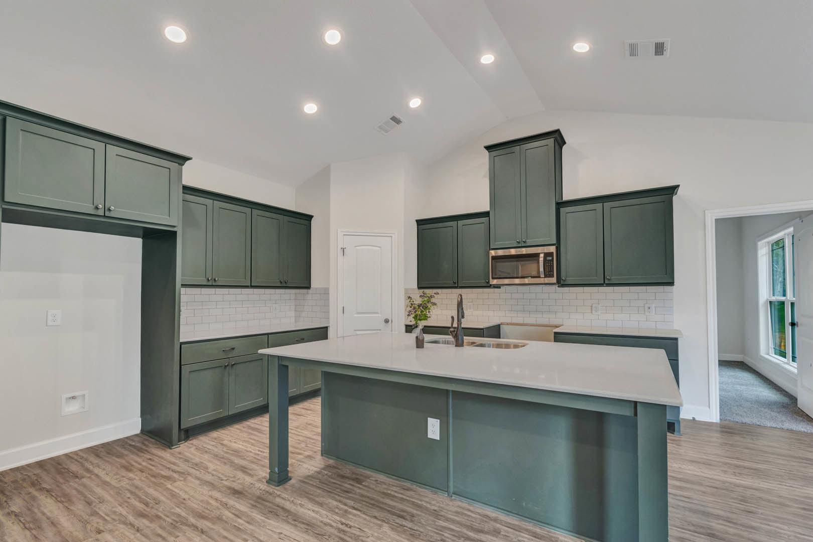 Spacious kitchen featuring green cabinetry, white countertops, large central island with built-in sink, tile backsplash, stainless steel microwave, and a vase with greenery
