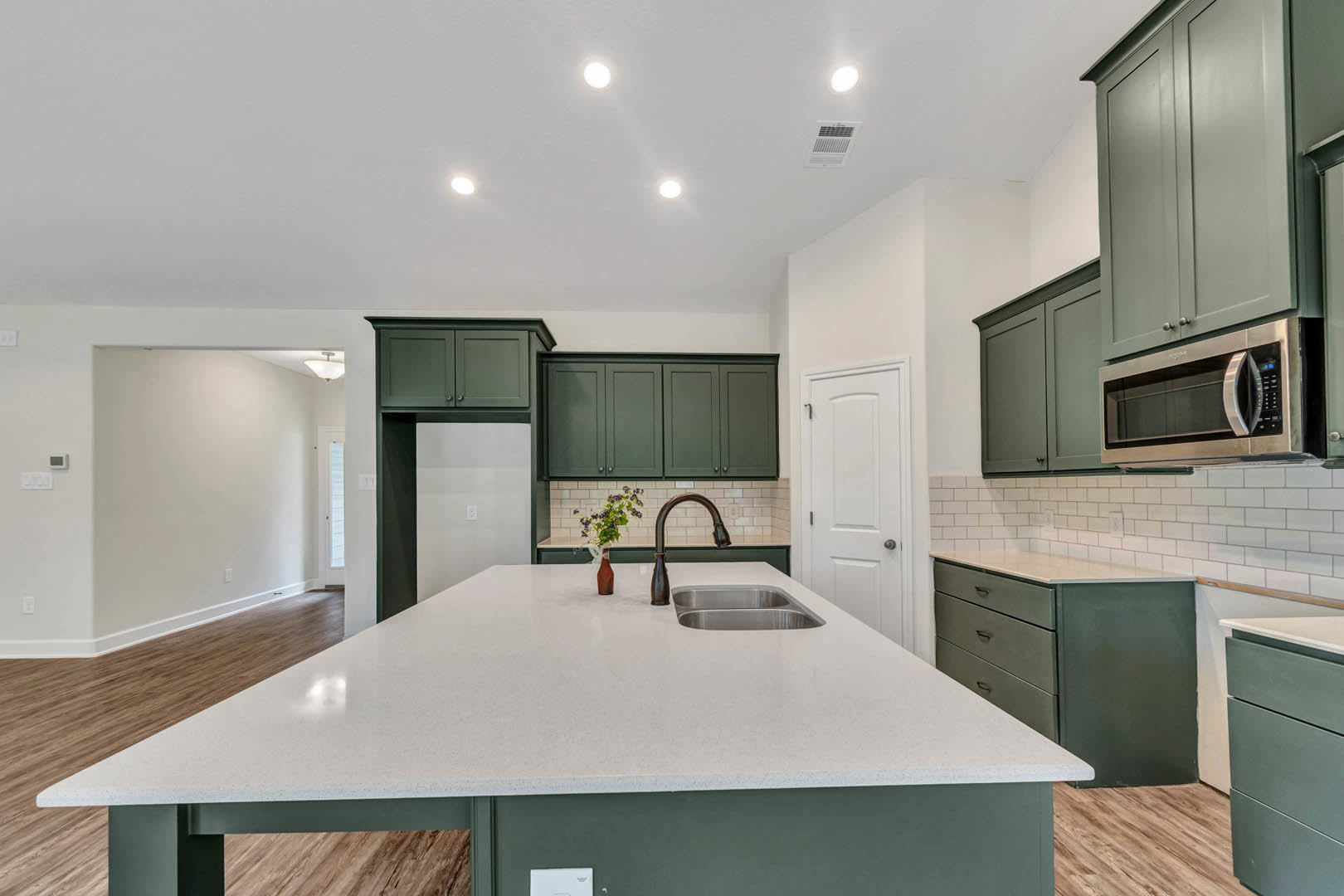 Green lower cabinets and white countertop in a kitchen with stainless steel microwave oven, white sink, vase of flowers, and visible drawer and wall outlet