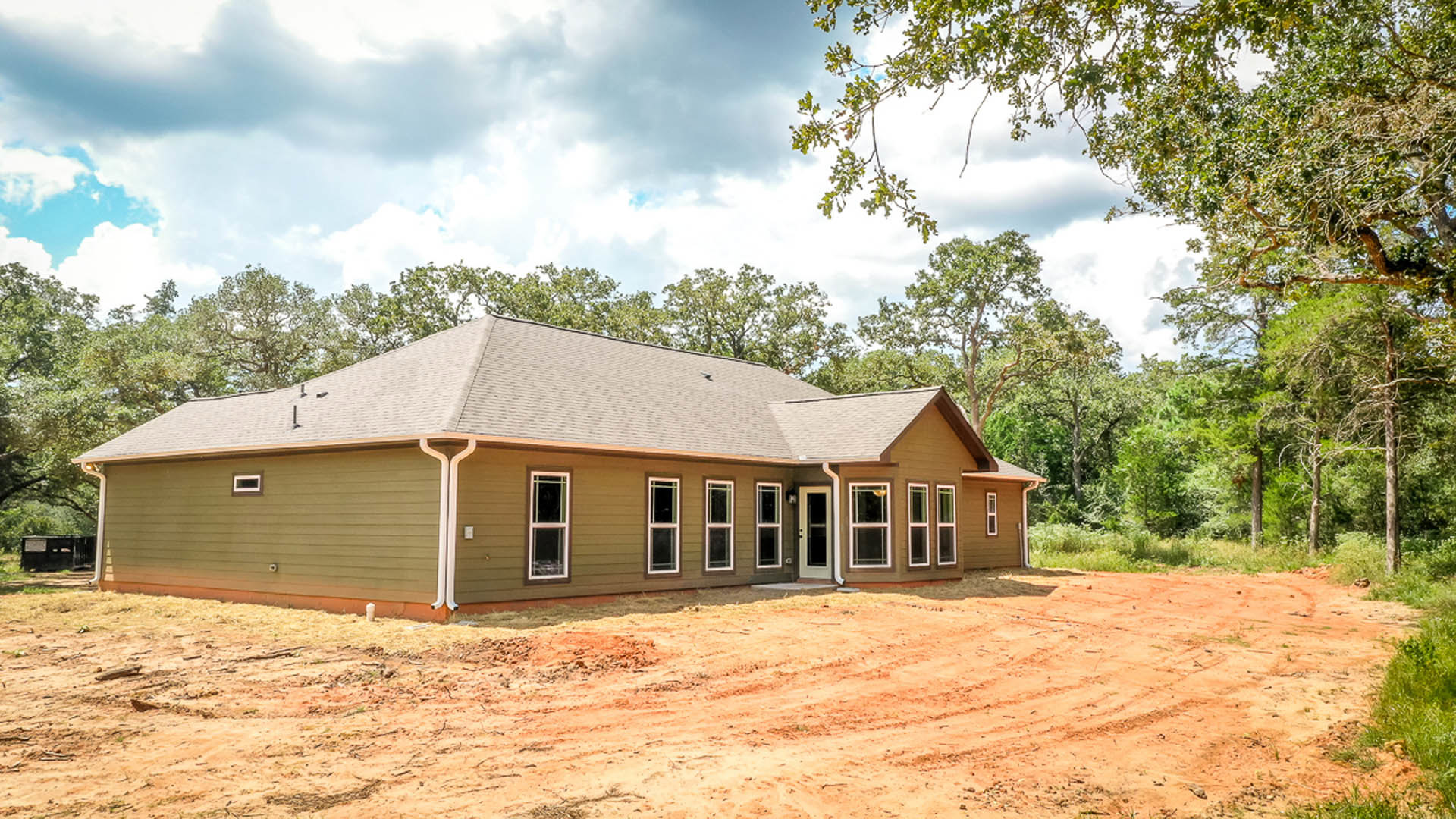 Partially built house with exposed framing, white and pink window frames, dirt yard, surrounded by trees under cloudy sky