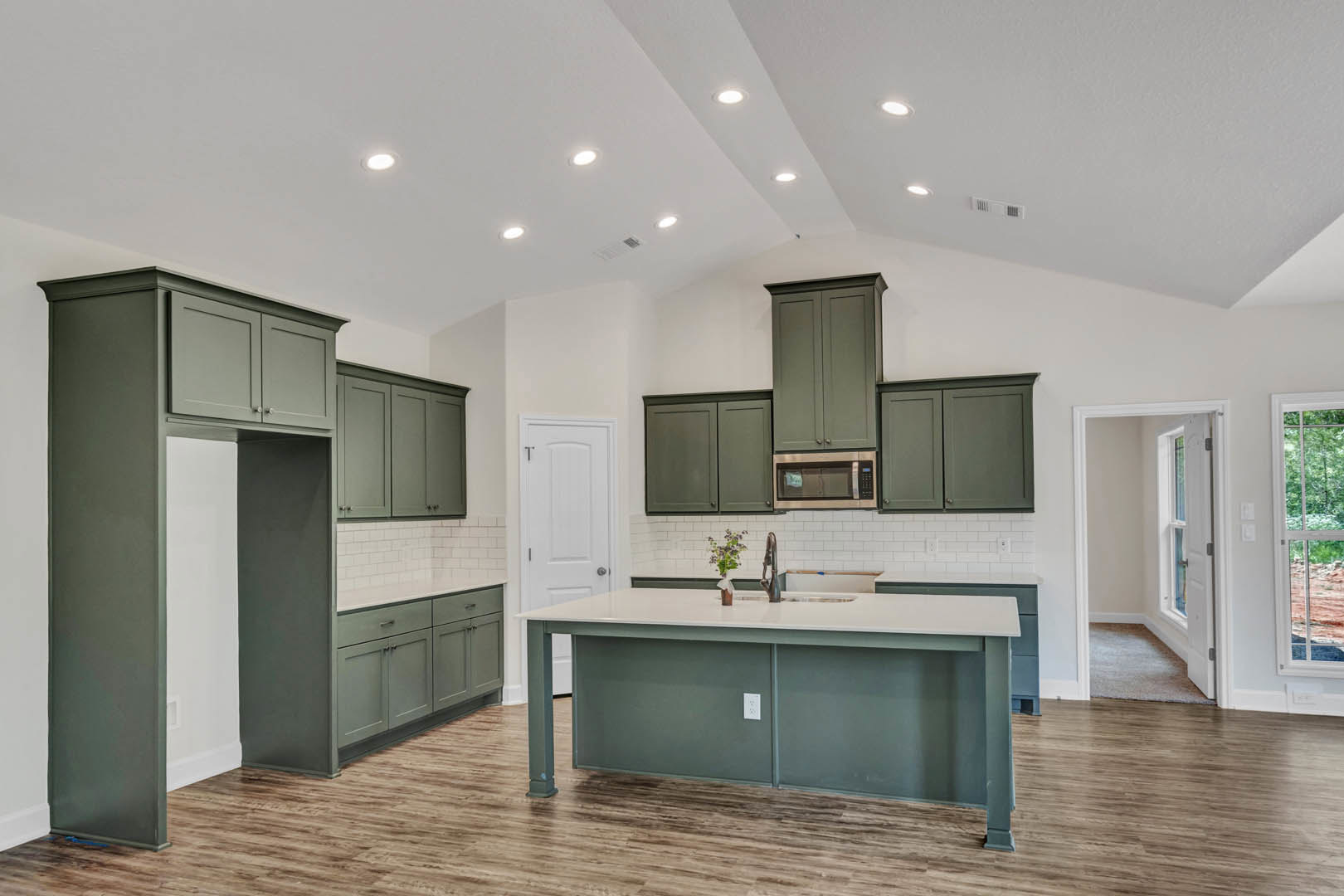 Spacious kitchen featuring a large central island with stone countertop, built-in sink, potted plant, white cabinetry, tile backsplash, and windows offering outdoor views and