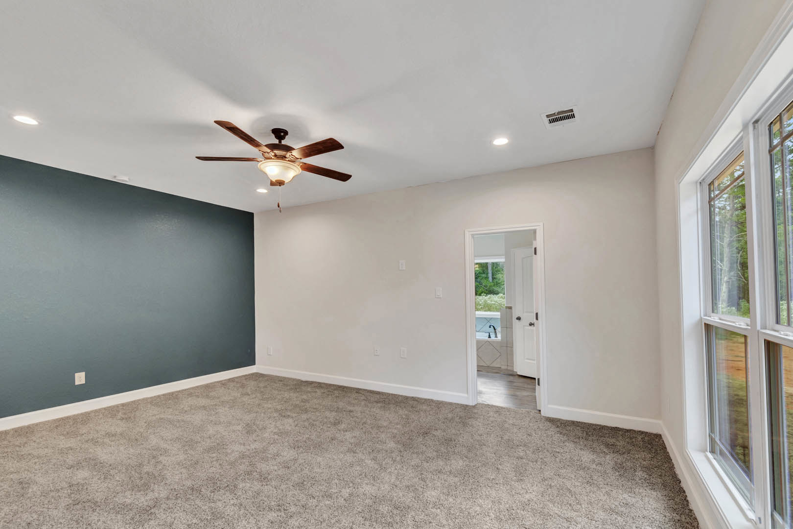 Carpeted bedroom with blue accent wall, white baseboards, ceiling fan with light fixture, door leading to bathroom featuring a window with tree views