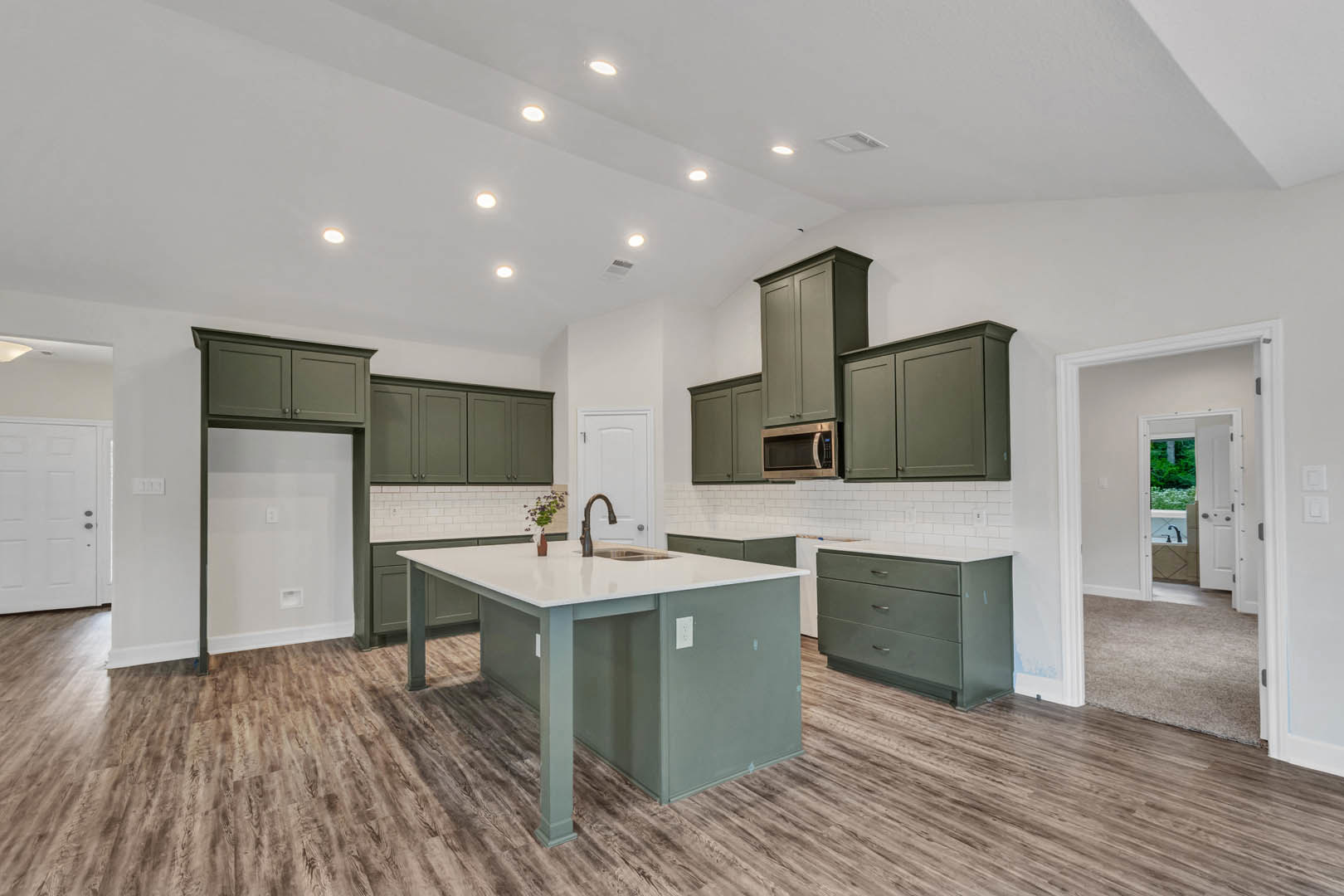 Spacious kitchen featuring a large central island with a built-in sink, light stone countertops, white cabinetry, and a vase atop the island; hardwood flooring and an open doorway