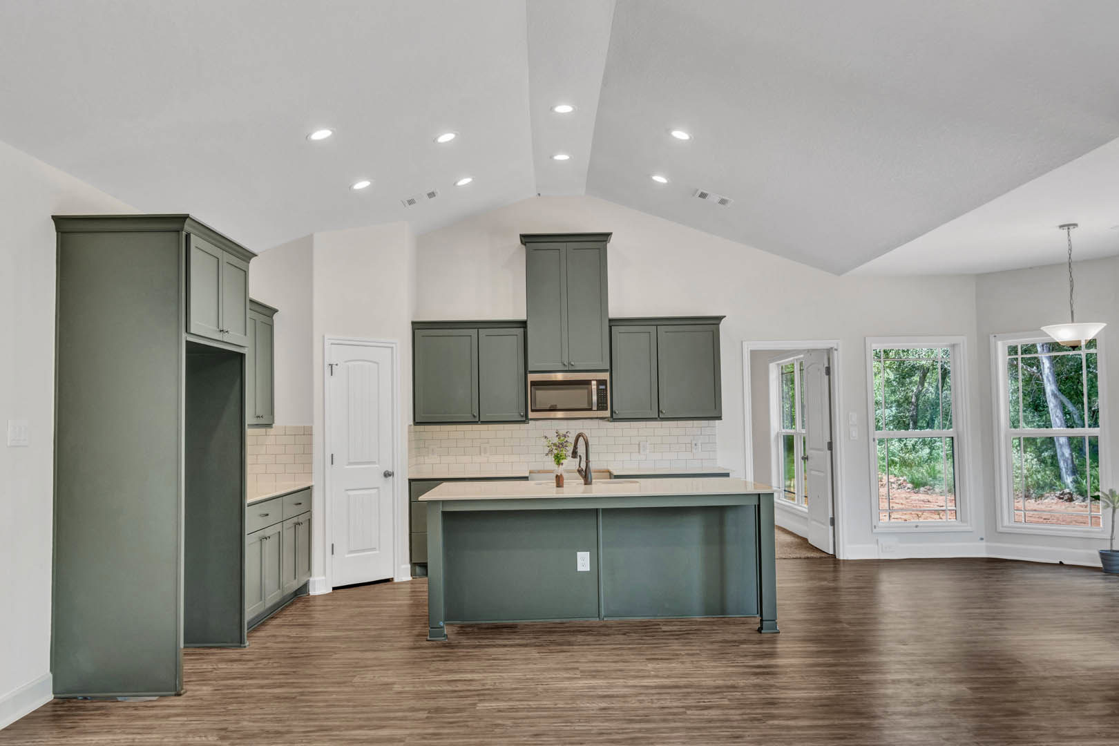 Spacious kitchen featuring a large central island with stone countertop, wood flooring, white cabinetry, stainless steel sink, built-in microwave, and a window overlooking trees.