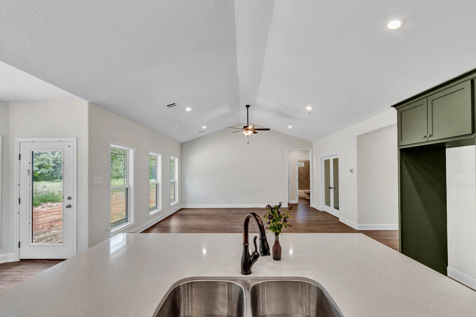 Open kitchen with wood flooring, white cabinetry, stainless steel sink and faucet, vase of purple flowers on the countertop, glass-paneled door, recessed ceiling light, black