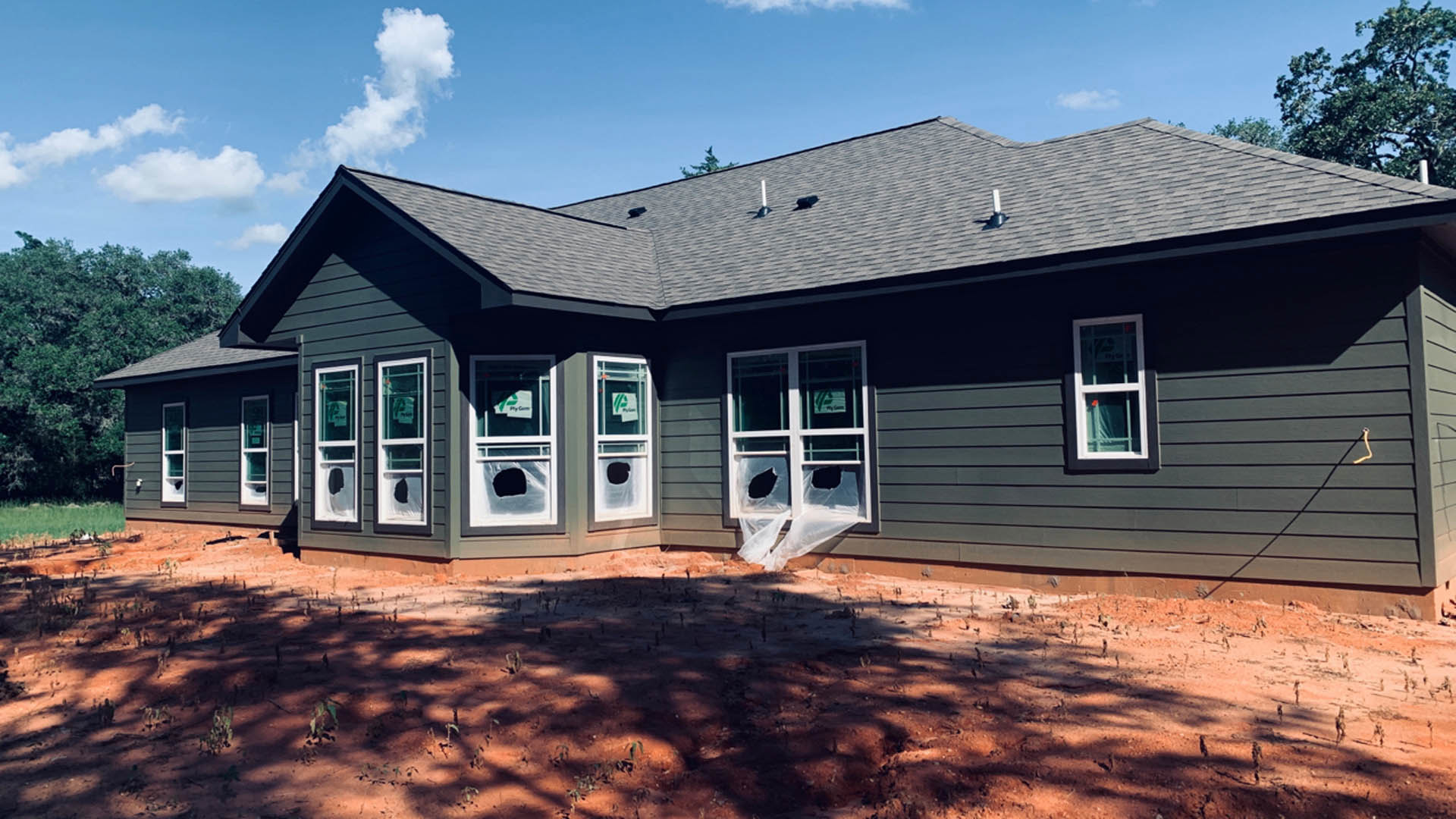Two-story house with white-framed windows, several panes broken, surrounded by a dirt field with small plants, trees in the background, cloudy sky overhead