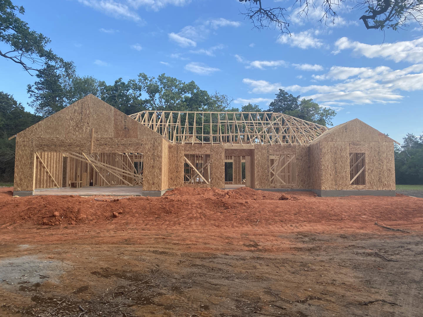 Wood-framed house under construction with exposed beams, dirt piles in the foreground, and mature trees under a blue sky with scattered clouds