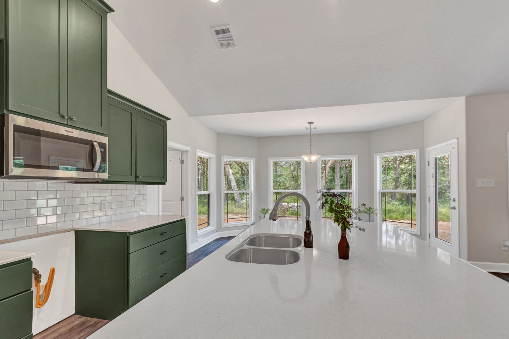 Green cabinetry kitchen with white countertops, stainless steel sink, built-in microwave, vase of flowers on counter, window view of tree trunk.