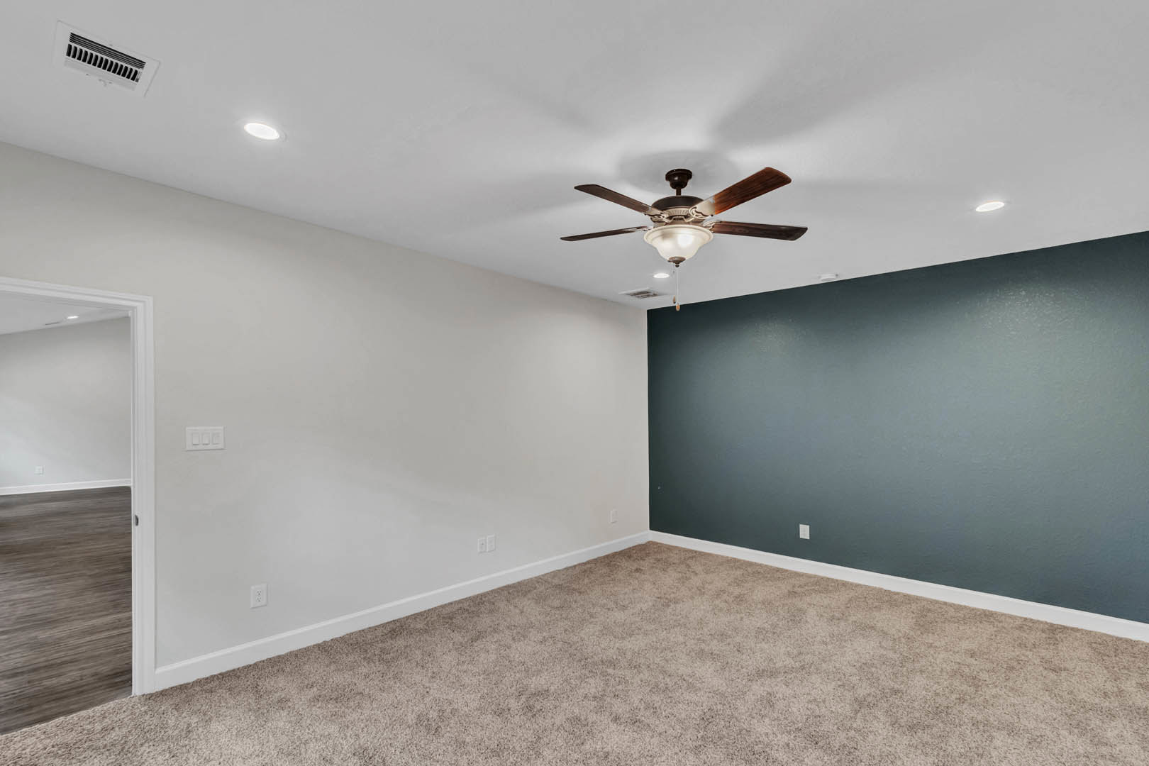 Ceiling fan with light fixture mounted above wood floor, carpeted walls, and plaster ceiling vent in custom home interior