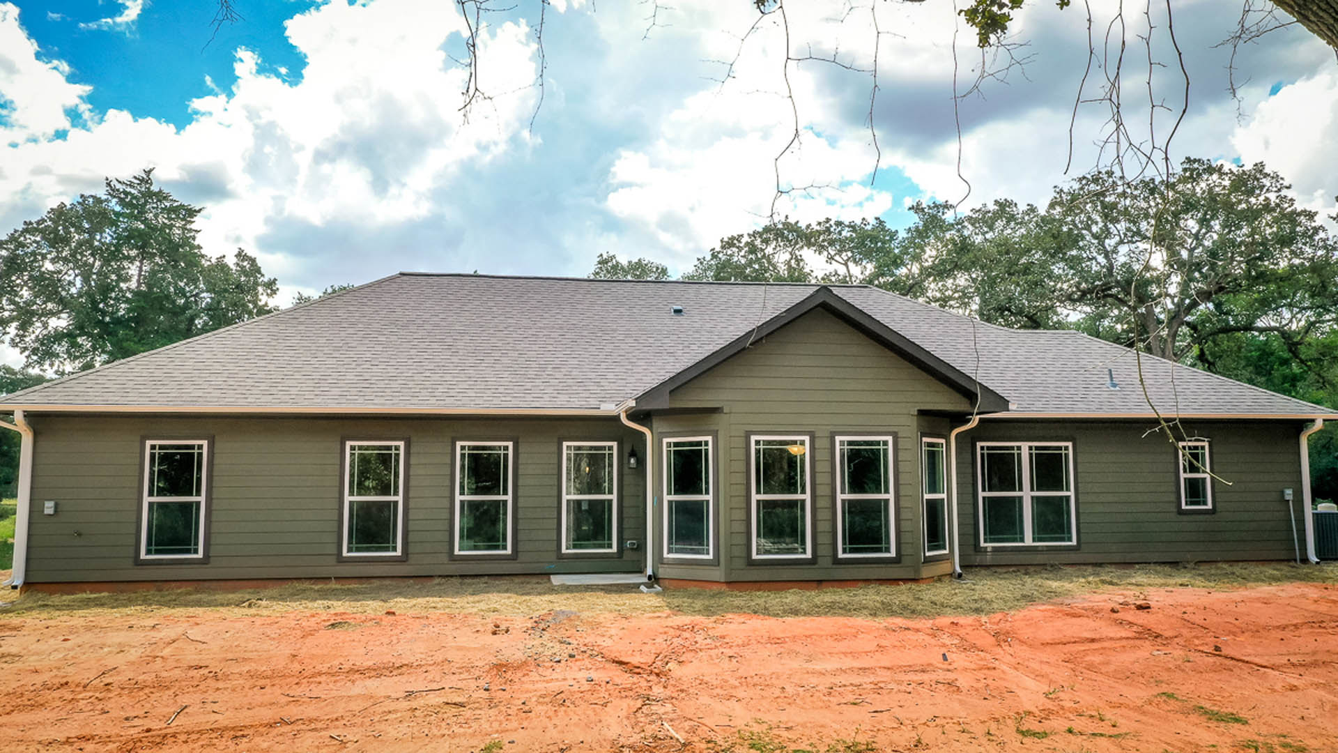 Partially built house with grey roof, white-framed window with broken glass, red dirt ground, surrounded by trees under cloudy sky