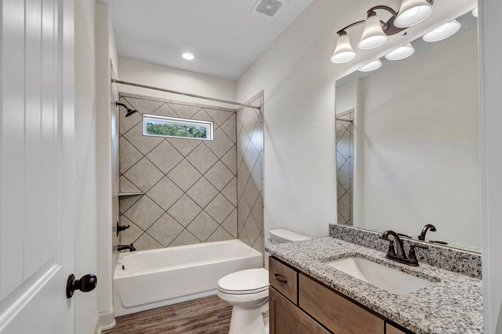 Bathroom with white tile walls, a rectangular sink and chrome faucet set in a stone countertop, white toilet, bathtub, large window overlooking trees, and a wall-mounted mirror