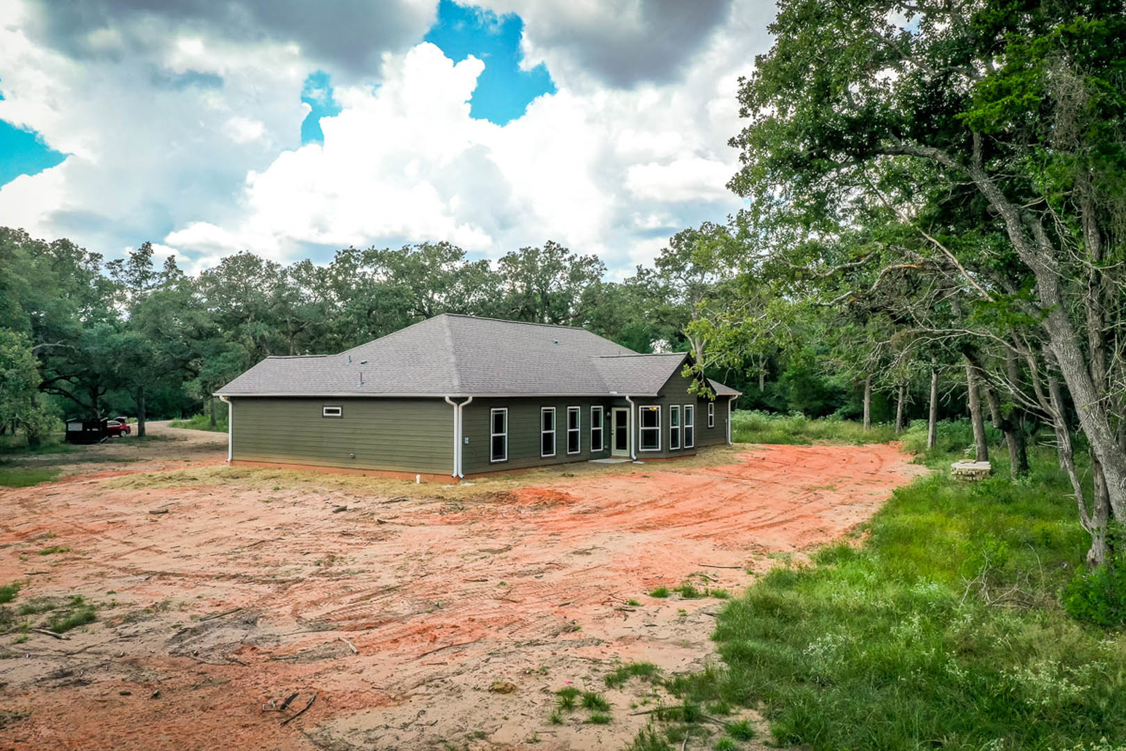 Partially built home with exposed framing and roof, surrounded by dirt lot and tall trees under blue sky with scattered clouds