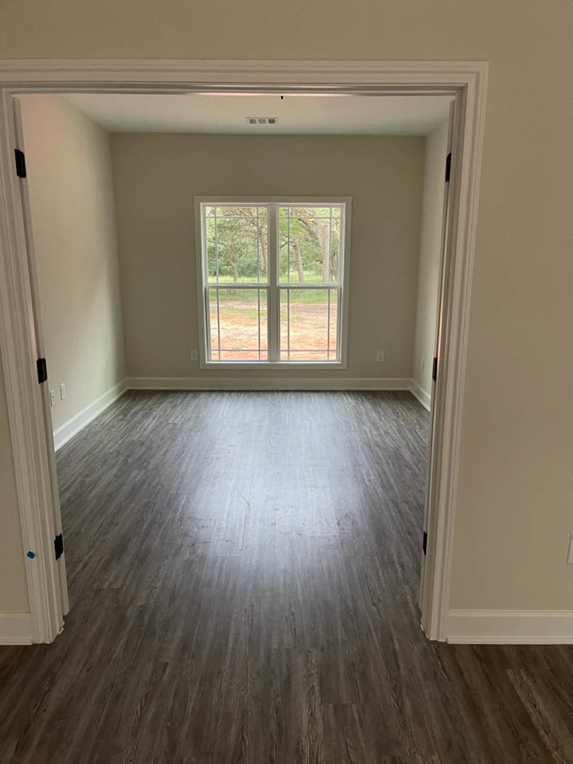 Sunlit room featuring wide-plank hardwood flooring, large window with tree views, white walls, and a close-up of a paneled door