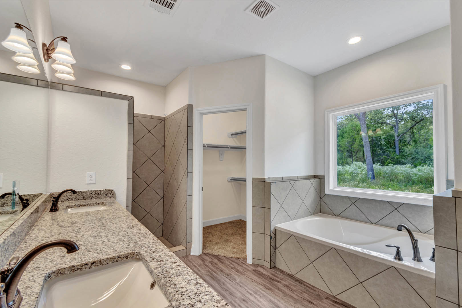 White freestanding bathtub with chrome faucet beside large window overlooking trees, modern sink with stone countertop, tile walls, round mirror, and contemporary lamp fixture
