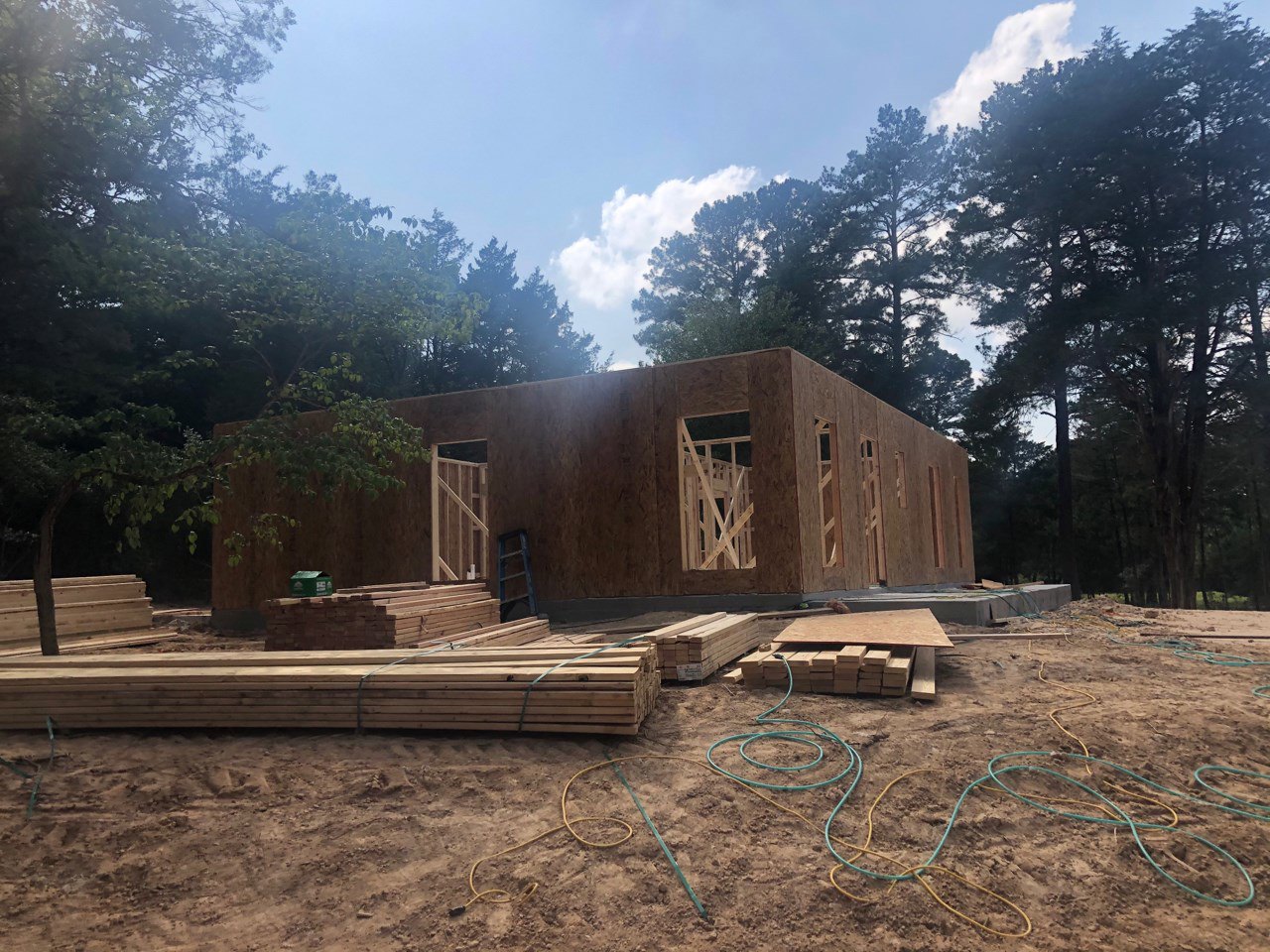 Wood-framed house under construction with exposed beams, stacked lumber, stairs, ladder, and trees in the background