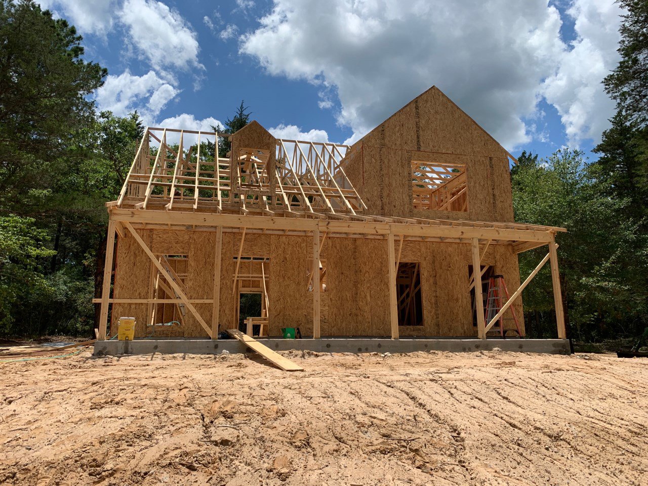 Framed house under construction with exposed wooden beams, dirt ground, scattered planks, and leafy trees in the background under a cloudy sky