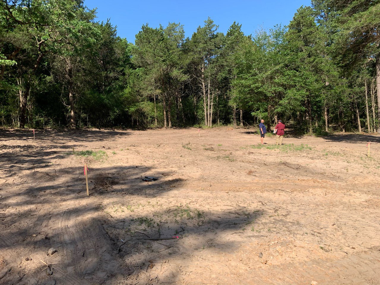Several people walking across a dirt clearing bordered by tall trees under a blue sky, with natural soil and sparse grass visible.