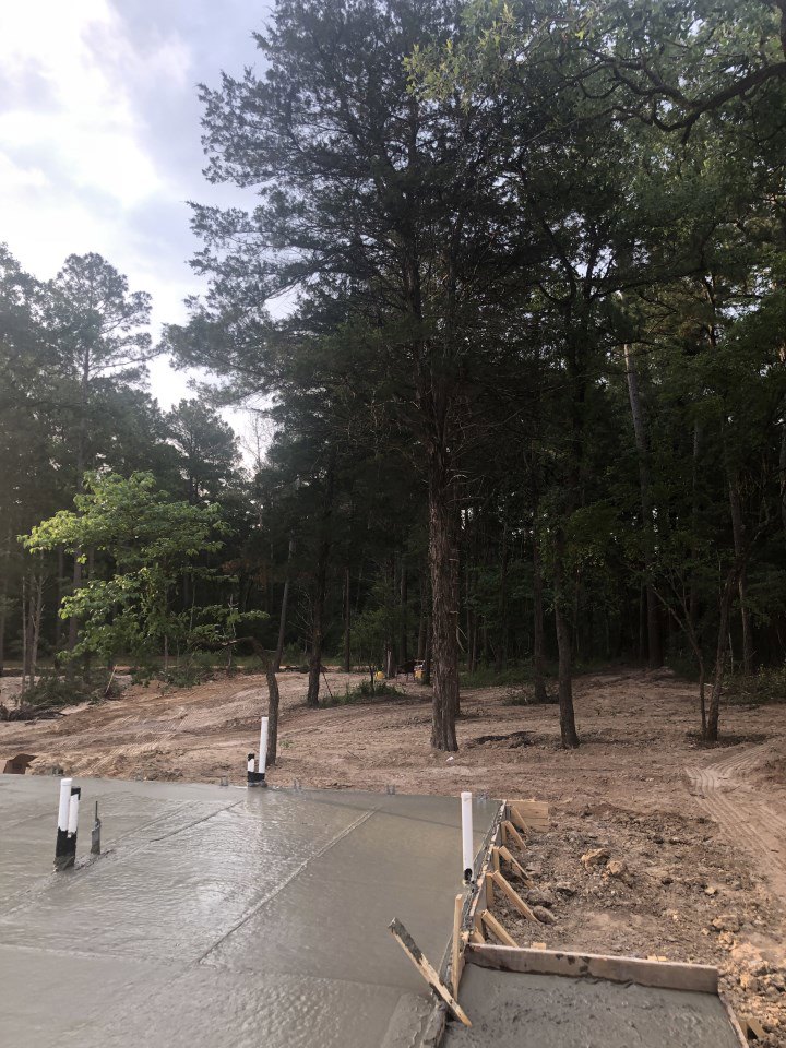 Freshly poured concrete slab surrounded by dense trees, wooden beams, and a vertical pole in a wooded outdoor setting
