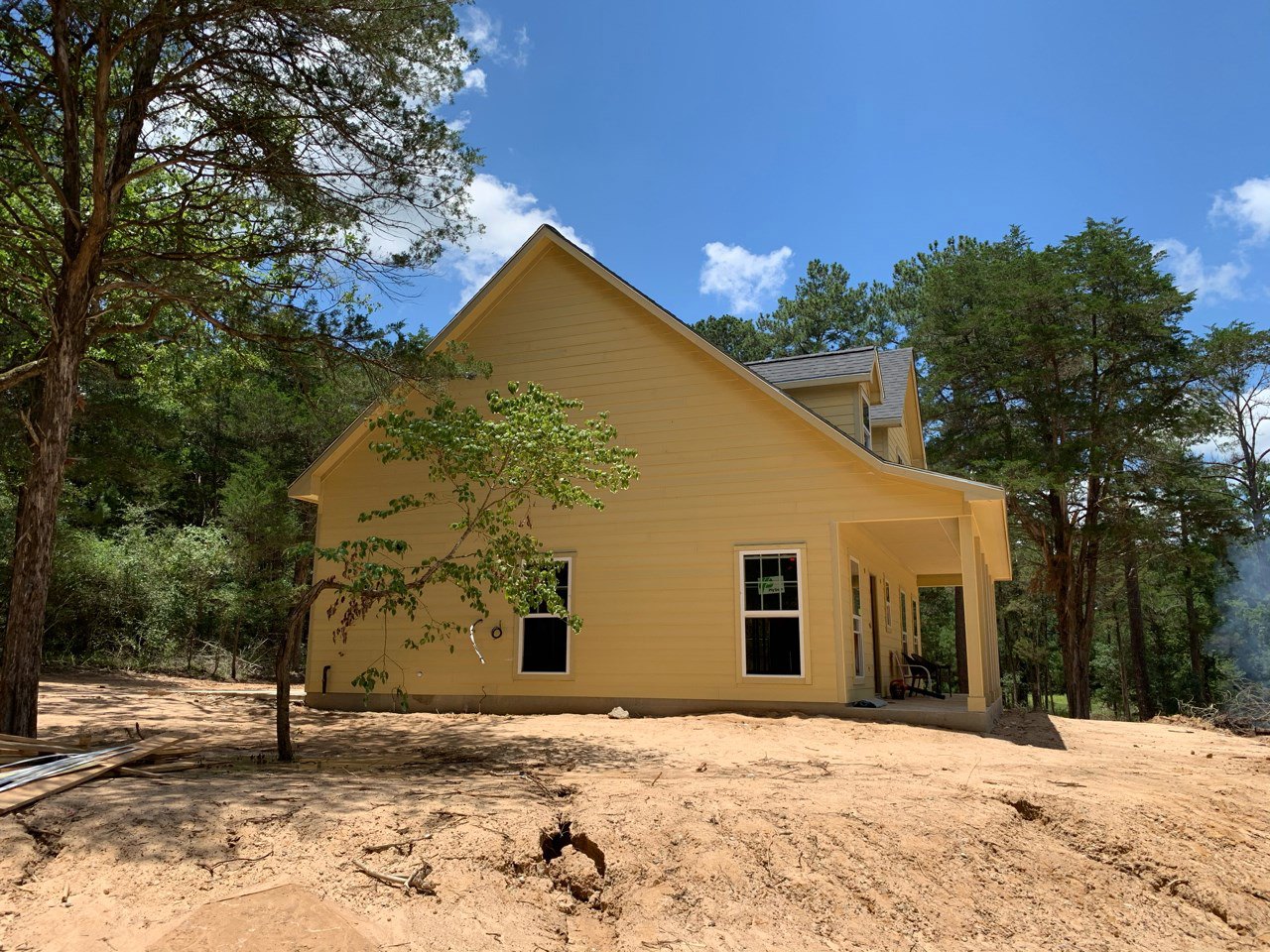 Yellow siding house with white trim, surrounded by mature trees, cloudy sky overhead, dirt patch in foreground, window displaying a sign, black planter with green foliage near