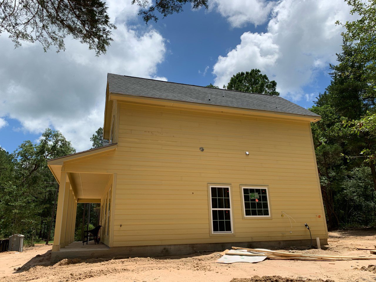 Yellow house with unfinished exterior, porch featuring a single chair, multi-pane window with sticker, pile of wood on bare ground, cloudy sky and trees in background.