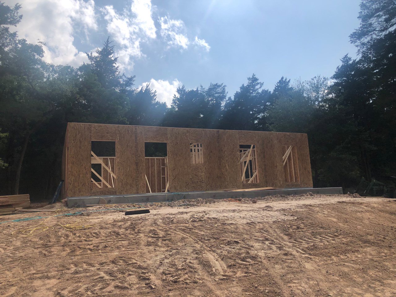 Wood-framed custom home under construction with exposed brick, large window opening, sandy ground with tire tracks, and trees in the background