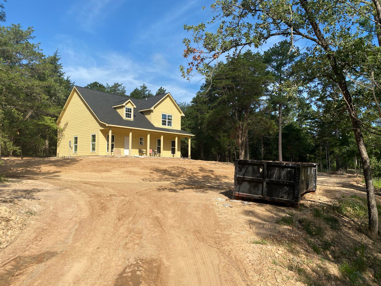 Yellow house with black roof, black dumpster with white paint in front, dirt road, leafless tree, cloudy sky