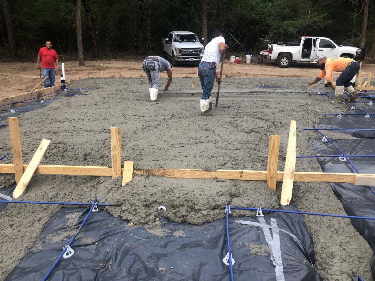 Crew pouring and leveling concrete slab foundation beside parked white pickup truck, surrounded by trees and dirt, with workers using tools and wearing hats and red shirts.
