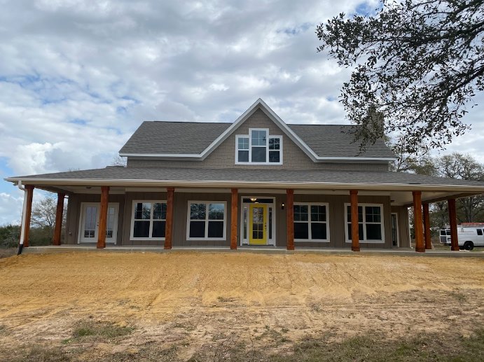 White siding home with multiple windows, yellow front door with glass panes, small porch, dirt and grass yard, leafy tree in foreground, partly cloudy sky.