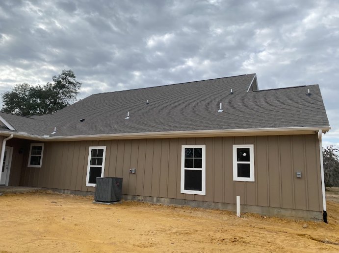 Wood-framed house under construction with exposed roof trusses, white-framed windows, and dirt lot beneath a cloudy sky