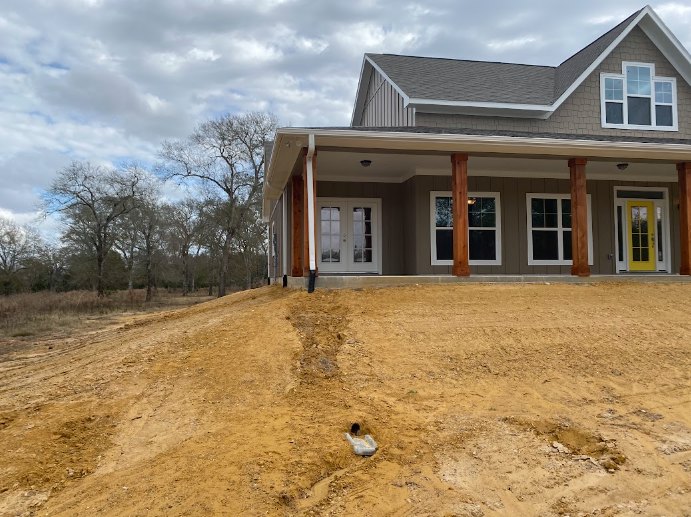 Modern house with yellow glass-paneled door, white double doors, wooden pillar, white-trimmed window, surrounded by dirt hill, trees, and cloudy sky