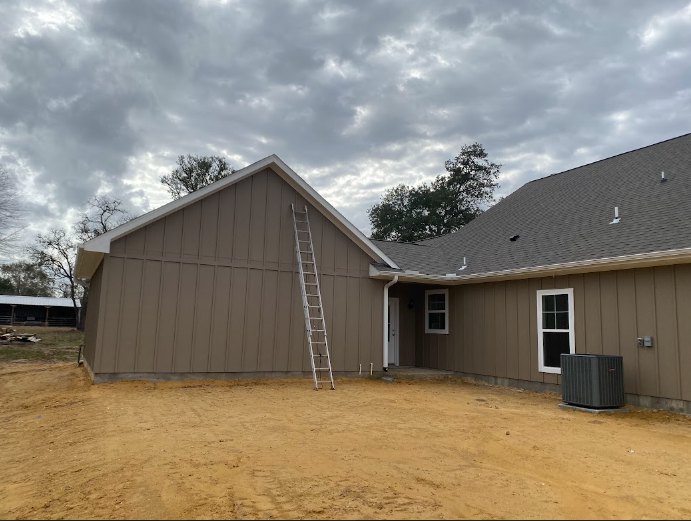 Partially built house with exposed framing, ladder leaning against exterior wall, dirt ground, black rectangular object on plywood, roof visible with trees in background under