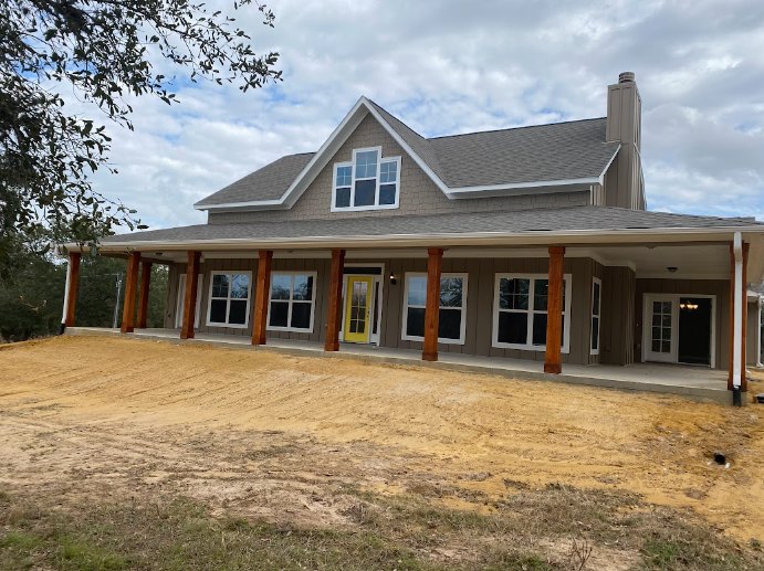 Two-story house under construction with exposed framing, dirt yard, surrounding trees, white door with glass panes, yellow accent door, multiple windows, cloudy sky overhead