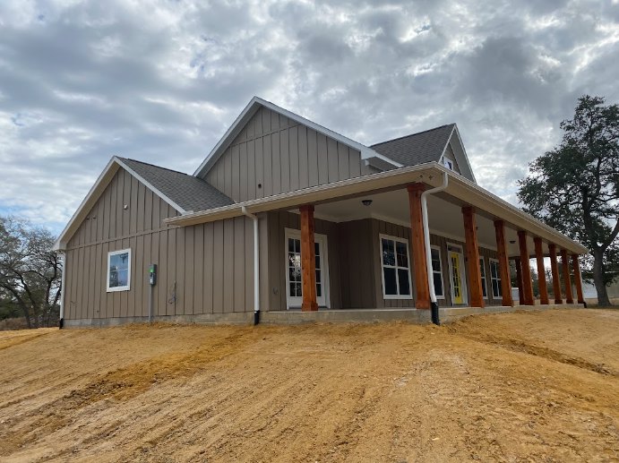 Wood-framed house under construction with exposed siding, covered porch, dirt hill in foreground, cloudy sky overhead, and scattered trees surrounding property