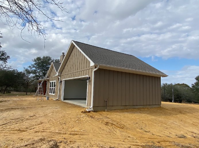 Two-story house under construction with exposed framing, white garage door, white entry door, ladder leaning against exterior, dirt patch in front, visible roofline, and