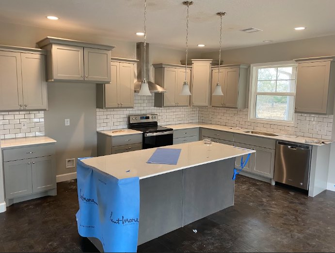 Spacious kitchen featuring a large central island with a blue towel draped over the side, white cabinetry, stainless steel stove and oven, built-in dishwasher, stone countertops